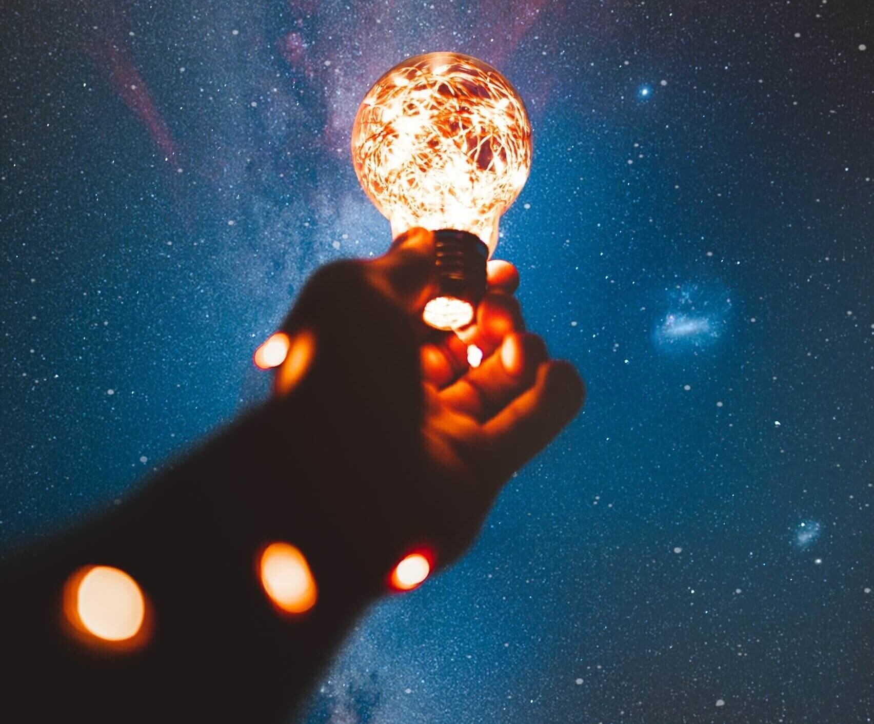 Image of a hand holding up a bright lightbulb against the night sky with stars